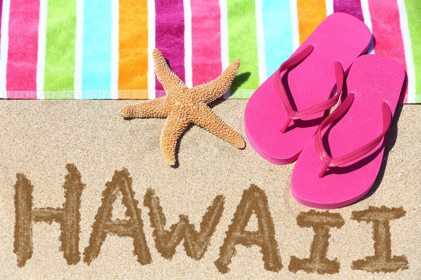Pink flip-flops, a starfish and a striped beach towel beside the word HAWAII written in sand: the basics of what to pack for Hawaii
